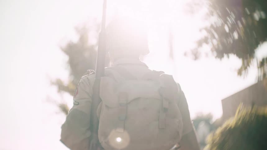 A soldier carrying a rifle and a backpack walks away from the camera into extreme sunlight creating a silhouette effect with heavy lens flare