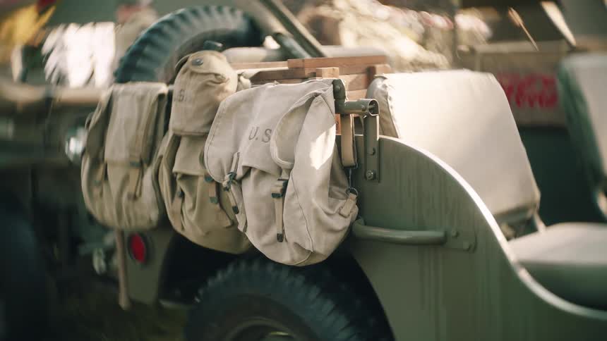 A close view of the back of a military jeep shows beige canvas bags and equipment secured over the rear wheel in a sunny outdoor setting