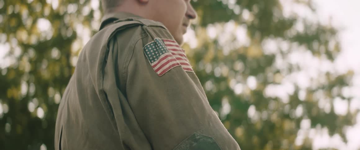 A soldier in a khaki uniform with an american flag patch on the sleeve stands outdoors in the bright sunlight