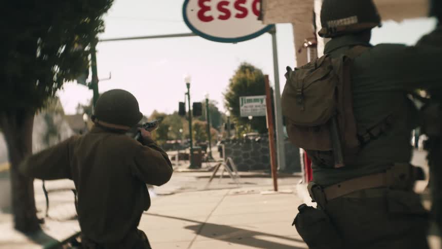 Military soldiers walk on the sidewalk in front of a gas station firing their guns