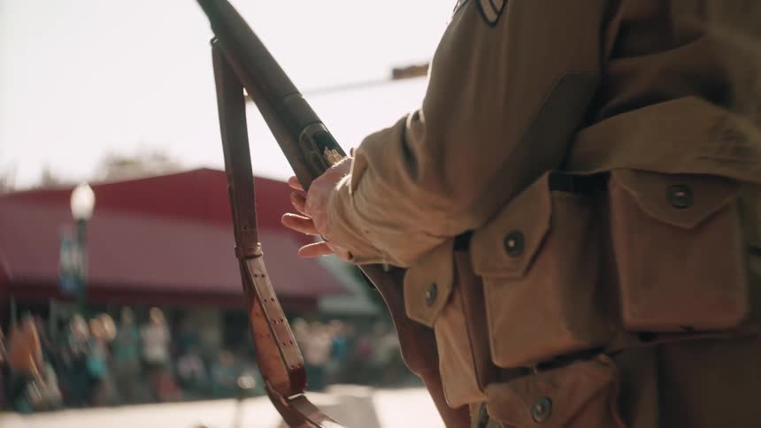 Military soldier stands on a street putting a bullet in their rifle as people watch in the distance