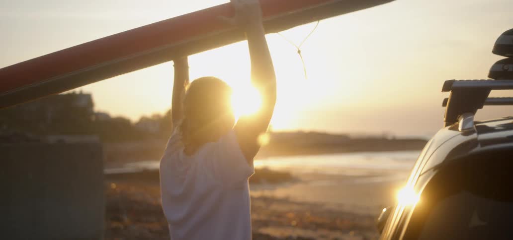 Man removing surfboard from top of car and carrying towards beach at sunset