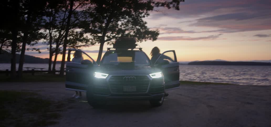 A man and woman park by a lake at sunset and get out of the car
