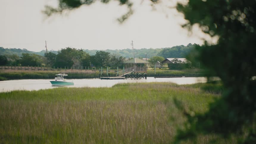 A small teal boat slowly moves across a peaceful river next to a wooden dock and green marsh grasses with a blurred tree in the foreground