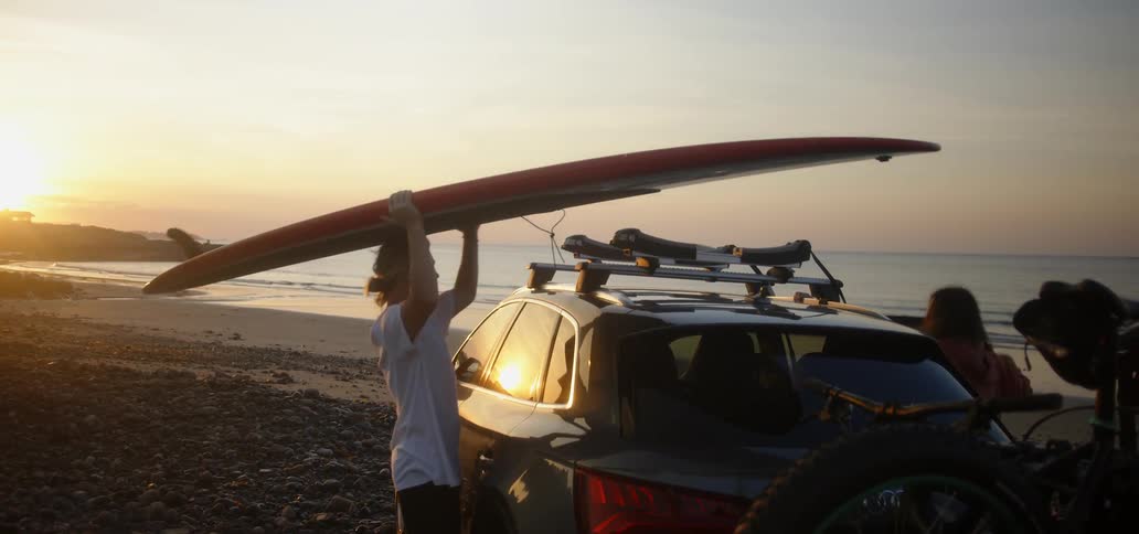 A man removes a surfboard from a car roof and walks towards the ocean at sunset