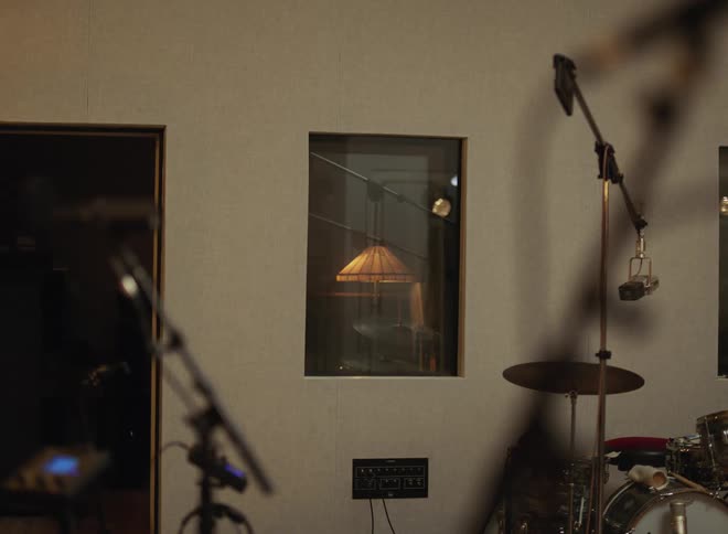 Recording studio with drum set suspended microphones and window into control room with equipment
