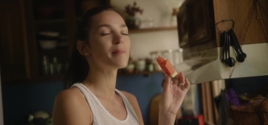 A woman stands in a kitchen eating some watermelon