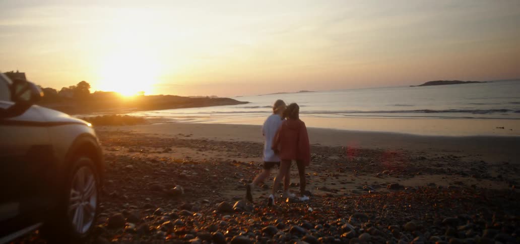 Man and woman walking from car down to ocean beach at sunset