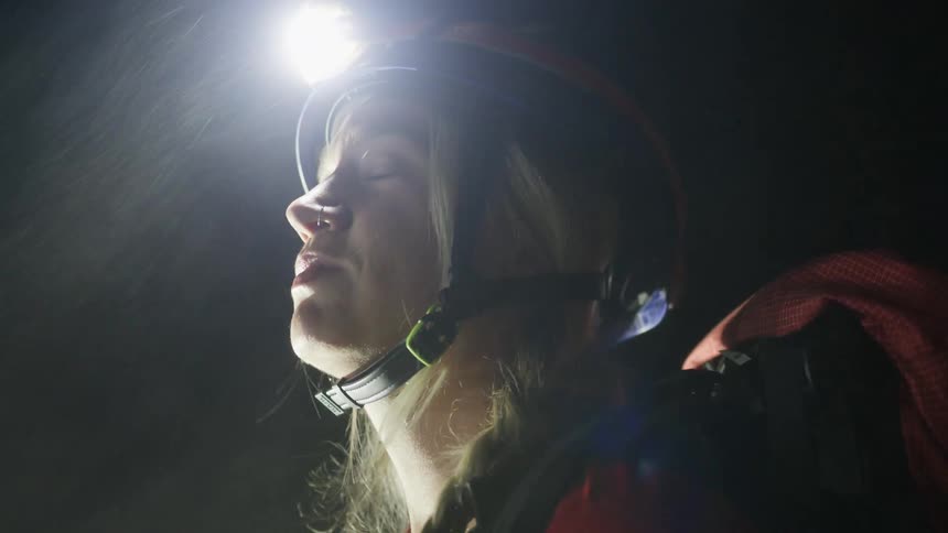 Woman wears a helmet with a headlamp and looks upward in a dark environment