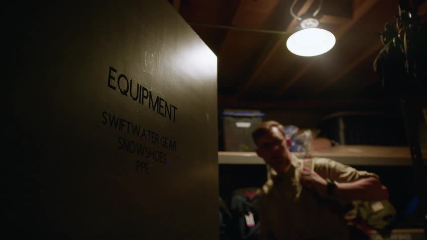 A man enters a dimly lit equipment closet grabs a backpack from the storage shelves and then turns to walk back out the doorway
