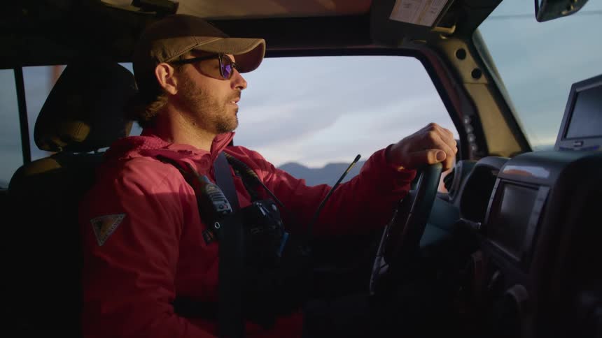 Person driving vehicle wearing red jacket cap and sunglasses in rural setting with cloudy sky