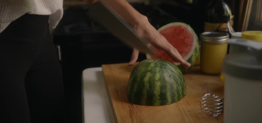 A person standing in a kitchen and cutting up a watermelon
