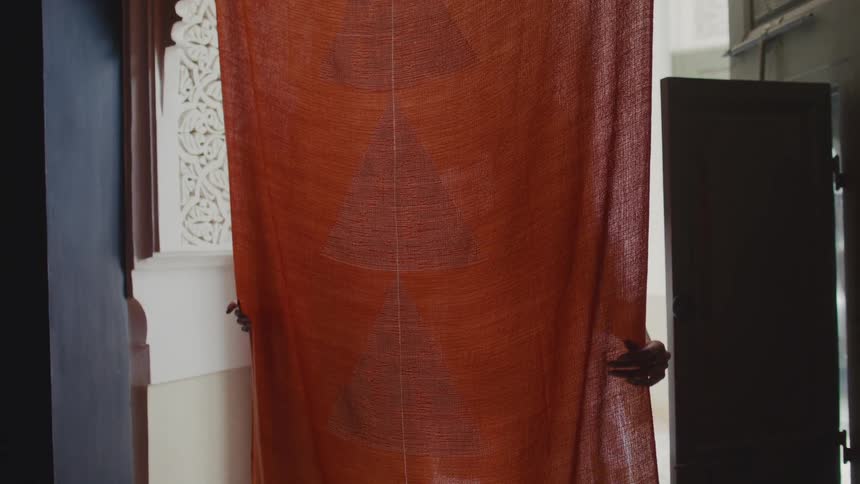 A pair of hands holds up a large translucent orangebrown fabric with a triangular pattern in a bright interior room flanked by ornate white carvings on the left and a dark wooden door on the right