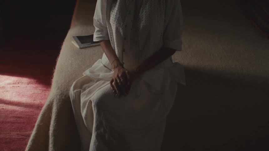 A young woman sitting on a bed in a softly lit bedroom wearing a white gown with a notebook beside her as daylight filters faintly through the window