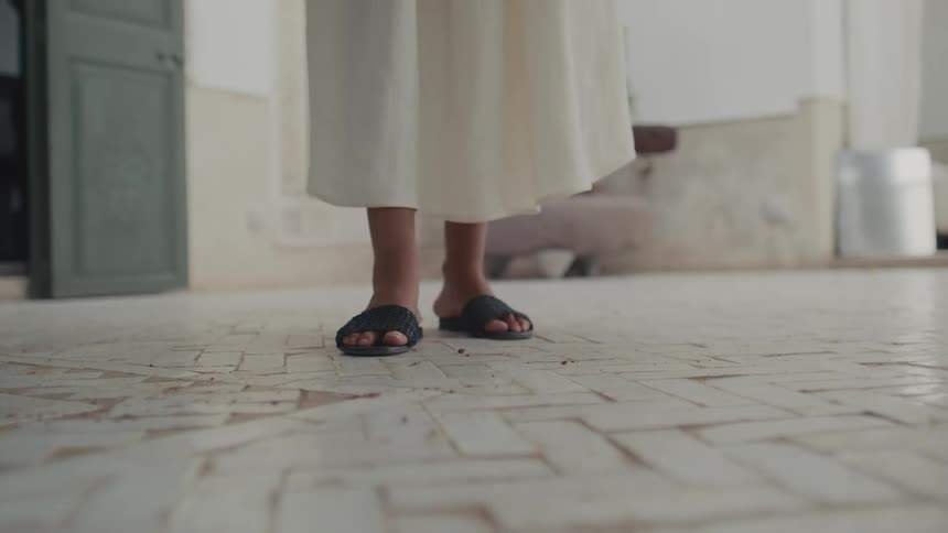 A woman standing outdoors on a front porch at midday wearing black slippers and a white gown on a tiled floor