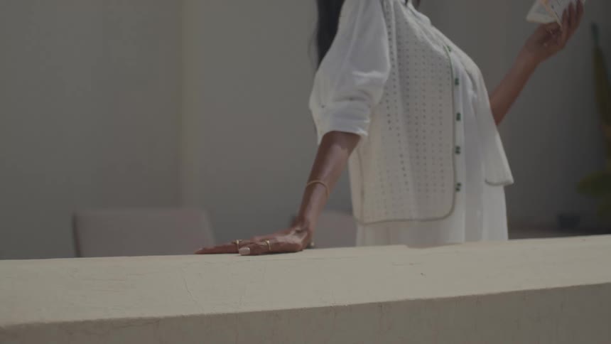 A woman in a white ensemble with gold jewelry leans her right hand on a pale wall or ledge while casually holding a small container in her left hand framed by a soft neutraltoned background