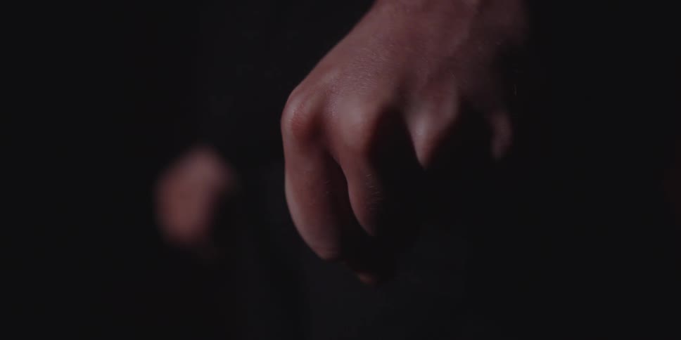 This is an extreme closeup shot focusing on a male handthe hand clenches slowly into a fist and then relaxes with prominent veins visible on the back of the hand against a dark featureless background