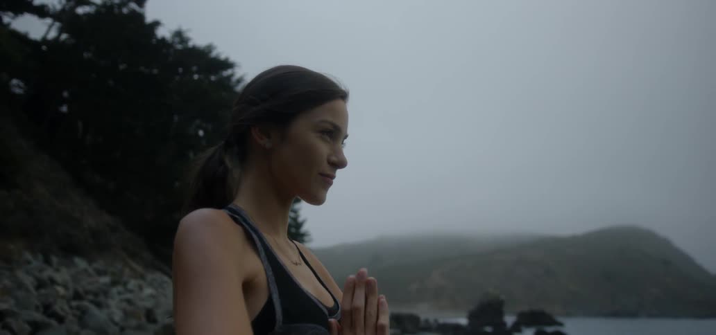 A woman practicing yoga outside on a cloudy day near a body of water and trees
