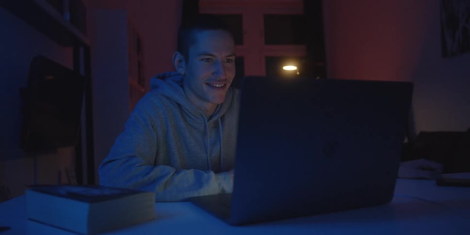 Man using a laptop at an indoor desk at night with a book beside it and blue ambient lighting
