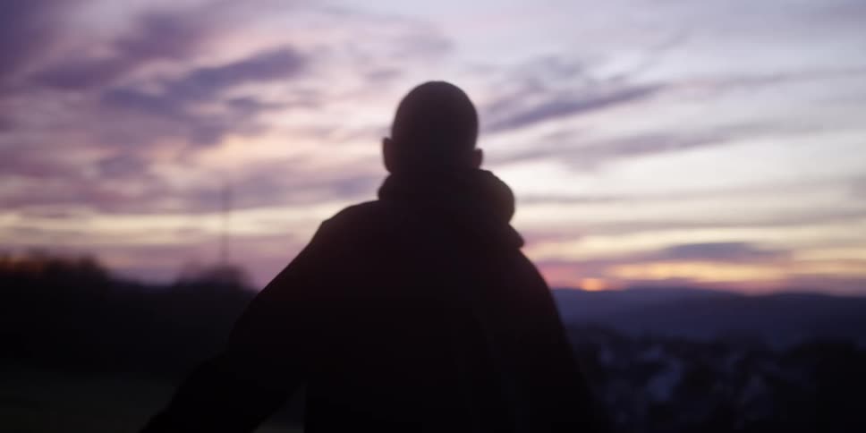A man seen in silhouette is standing outdoors with a colorful sunset visible in the backgroundhe turns his head looks up at the sky and then turns his back to the camera walking away slightly