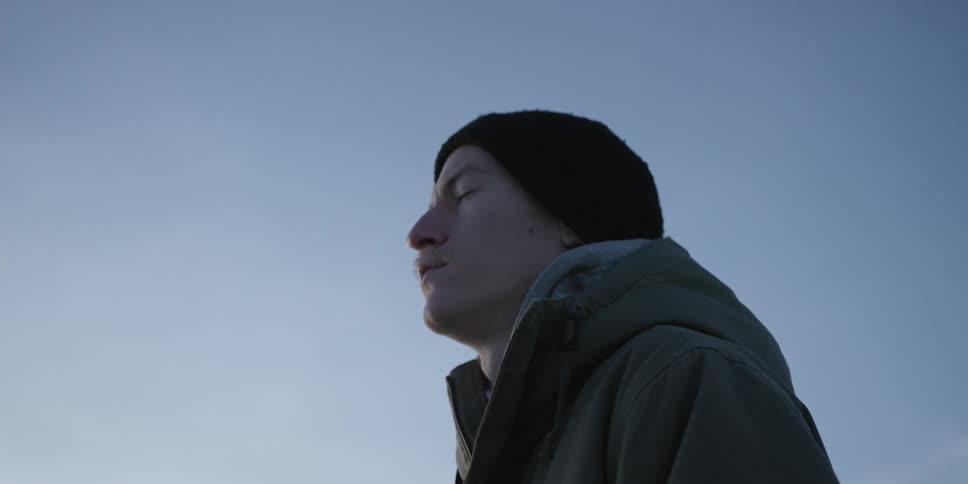 Person in profile wearing a black beanie and green jacket eyes closed facing the sky