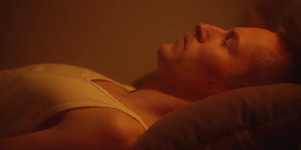 A closeup shot of a persons upper body lying on a pillow with their eyes closed and their face in profilethe scene is dimly lit with a strong warm orangeamber light giving the footage a cozy or intimate atmosphere