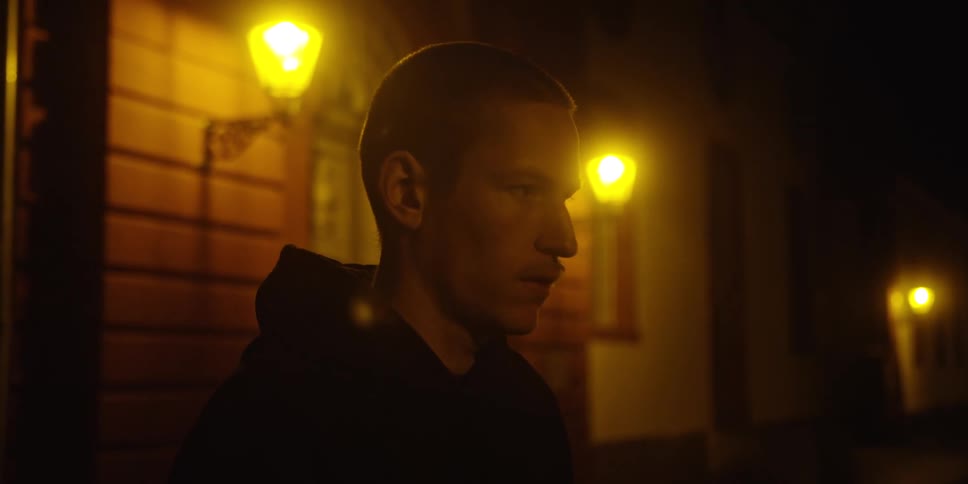 Man in profile on a street at night lit by yellow street lamps in the background