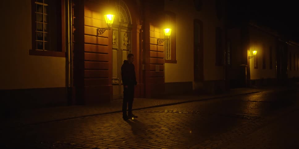 This is a wide shot of a young man standing on a cobblestone street at night illuminated by yellow light from decorative wall lanterns on the surrounding buildingsthe architecture suggests a historic european setting with the man standing near a larg
