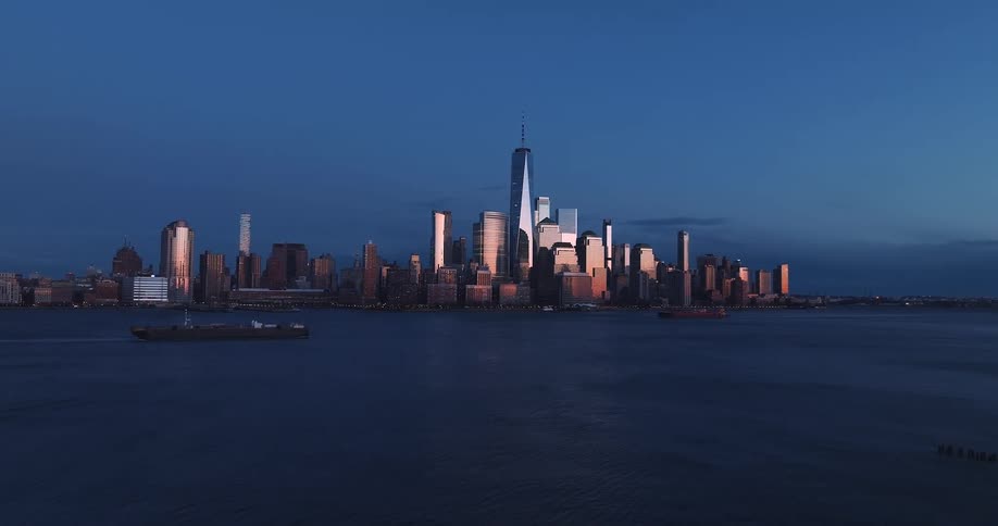A city skyline sits under a deep blue dusk sky as the setting sun casts a bright golden glow across the glass facades of the buildings and surrounding skyscrapers while ships move slowly across the river