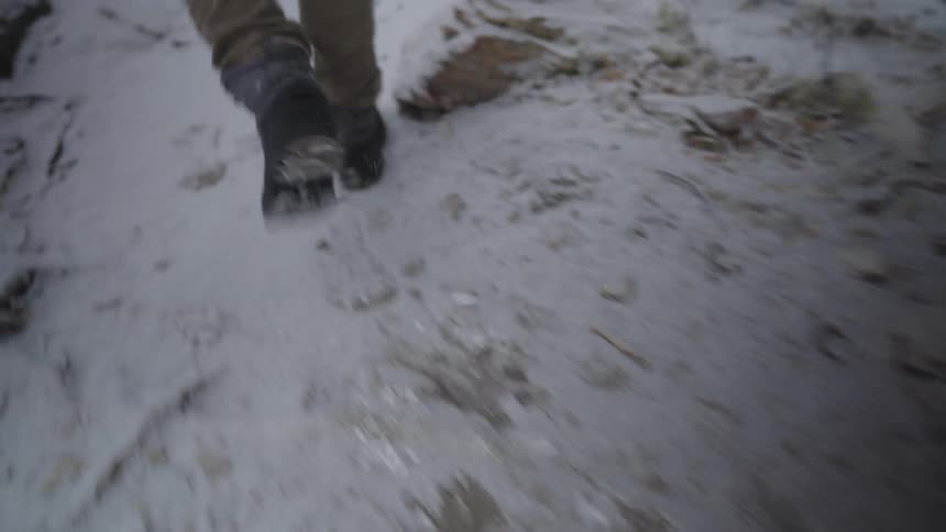 A close view from behind of a person wearing tan pants and gray boots walking across a snowy and rocky ground during winter