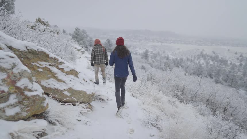 A man and a woman wearing winter jackets and hats walk away from the camera along a snowy mountain trail overlooking a foggy valley