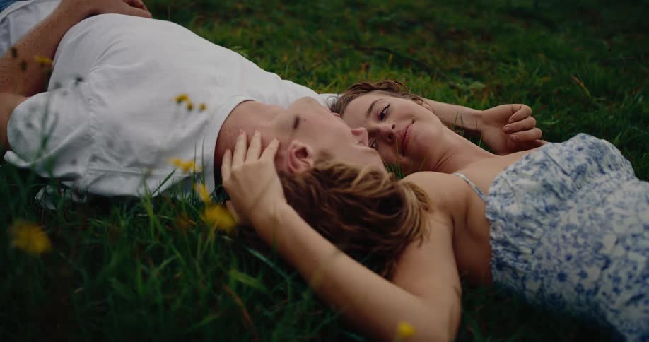 A young couple lies intimately together in lush green grass scattered with yellow wildflowers gazing at each other tenderly