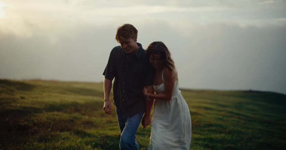 A young couple holding hands as they walk up a grassy hill at sunset warm light stretching their silhouettes across the slope