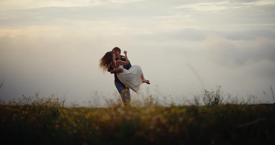 In a sweeping cinematic moment at dusk a man joyfully spins a woman in his arms across a vast grassy field as the soft evening light glows behind them