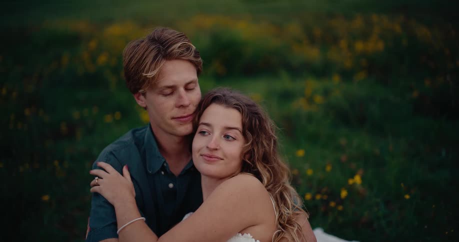 A young man and woman lie in a field of green grass and yellow wildflowers smiling at each other while the woman tenderly strokes the mans arms during an intimate moment outdoors