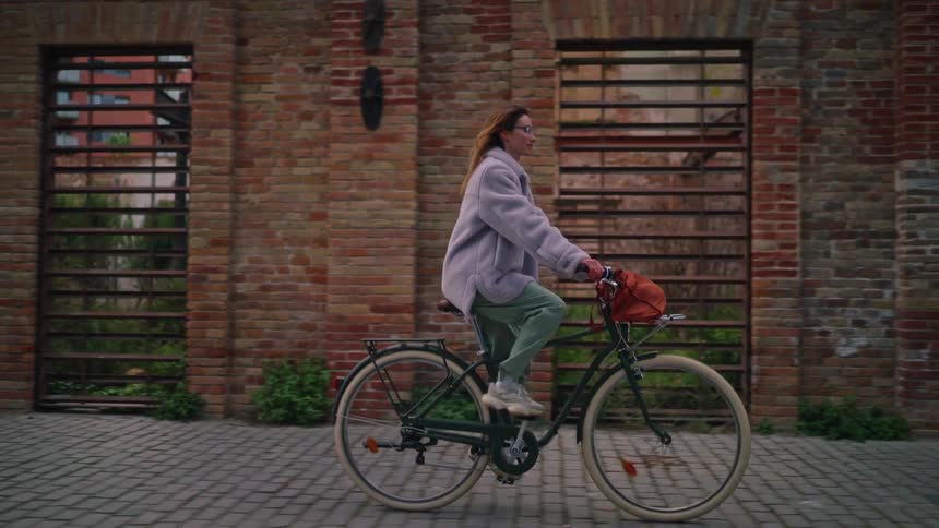 A woman in a lilac fleece jacket and glasses rides a vintagestyle bicycle past an old brick buildingthe camera follows her in a sideprofile tracking shot as she smiles and looks ahead toward the urban landscape