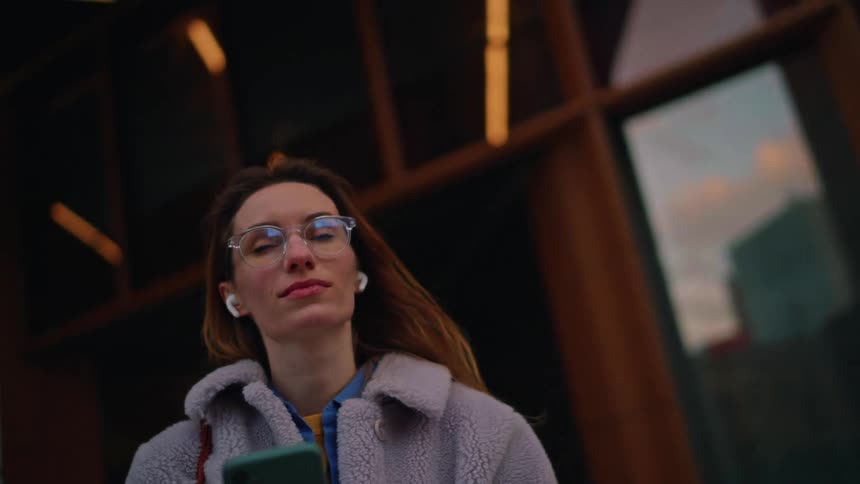 A young woman wearing glasses earbuds and a fluffy lightgray coat stands outdoors in an urban setting at dusk looking down and typing on her smartphone
