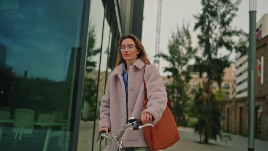 A stylish woman in a pink jacket rides her bicycle down a city sidewalk her reflection gliding across the large glass windows of a modern building beside her