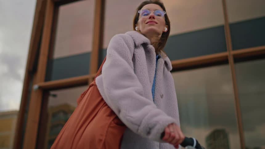 A woman in a fuzzy lilac coat and glasses walks alongside a modern building while pushing a bicycle she wears white earbuds and looks around at her urban surroundings under a cloudy sky