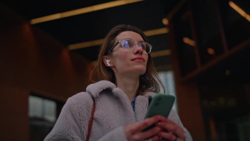 A woman in glasses looks up with a thoughtful expression as she walks past a modern building holding her smartphone while the camera circles around her