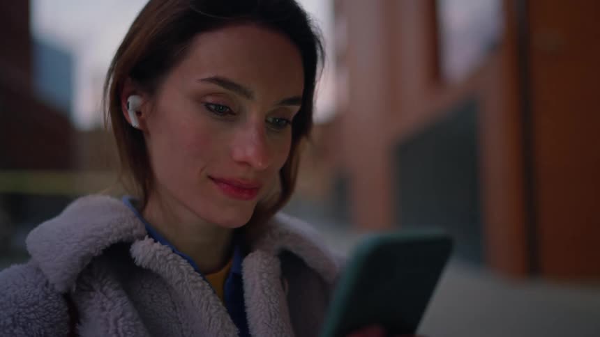 Woman using a smartphone with wireless earbuds in an urban setting at dusk