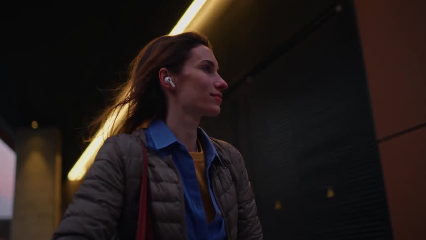 A woman with flowing hair rides a bicycle past a modern darkpaneled building at dusk wearing wireless earbuds a light jacket and a blue scarf while carrying a large rustcolored shoulder bag