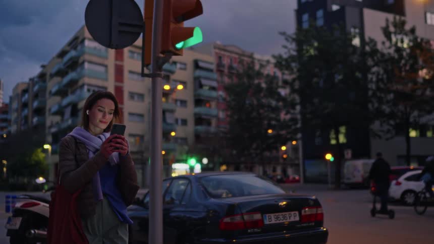 A young woman walks along a city sidewalk while texting on her smartphone at dusk