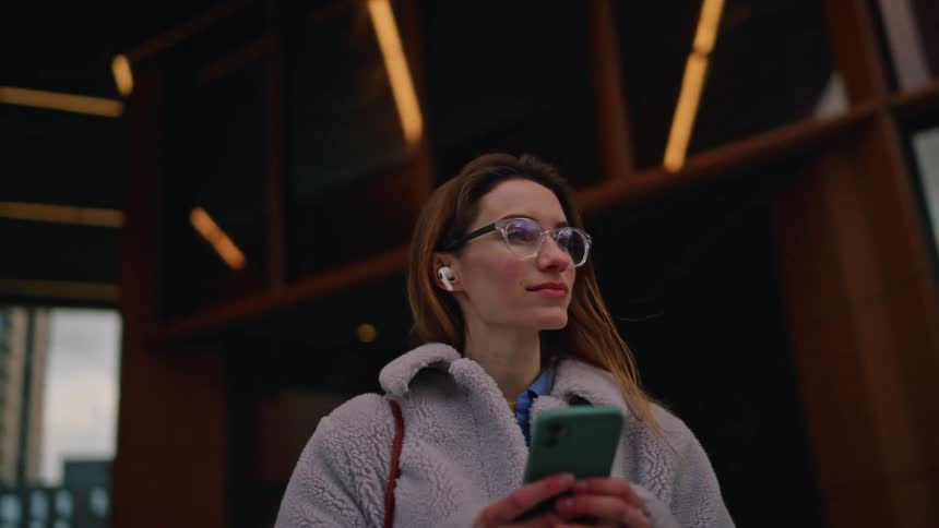 A woman looks at her phone while standing along a city sidewalk as the view orbits under an overcast sky with a modern building in the background