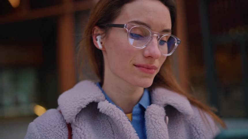 Woman wearing glasses and wireless earbuds while interacting with a smartphone in an urban setting