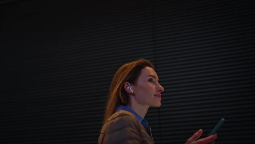 A woman walks alongside a dark textured wall at night while holding a smartphone and wearing white earbuds she wears a quilted brown jacket over a blue shirt and glances forward with a subtle calm smile