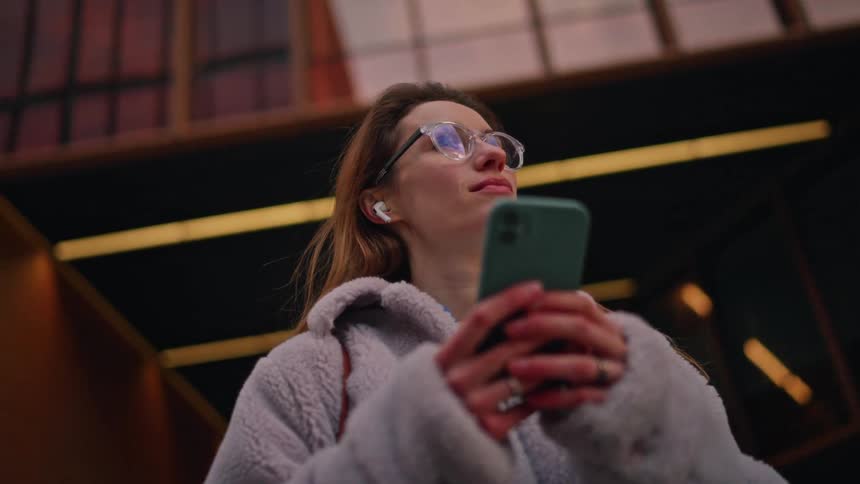 Woman using a smartphone with wireless earbuds in an urban space beneath a modern architectural structure