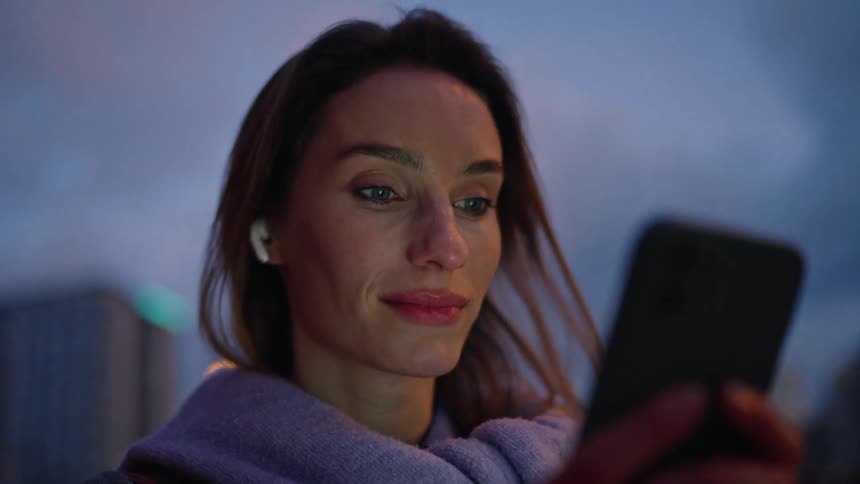A woman smiles while using her smartphone outside in the evening