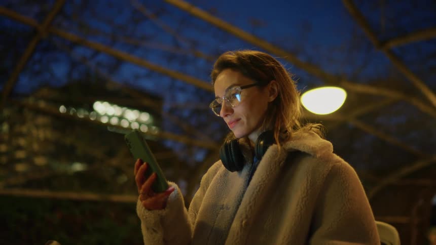 Under a dark blue evening sky a woman in a thick beige coat and glasses stands outdoors beneath a metal canopy holding up her smartphone and looking at the screen with a slight smile