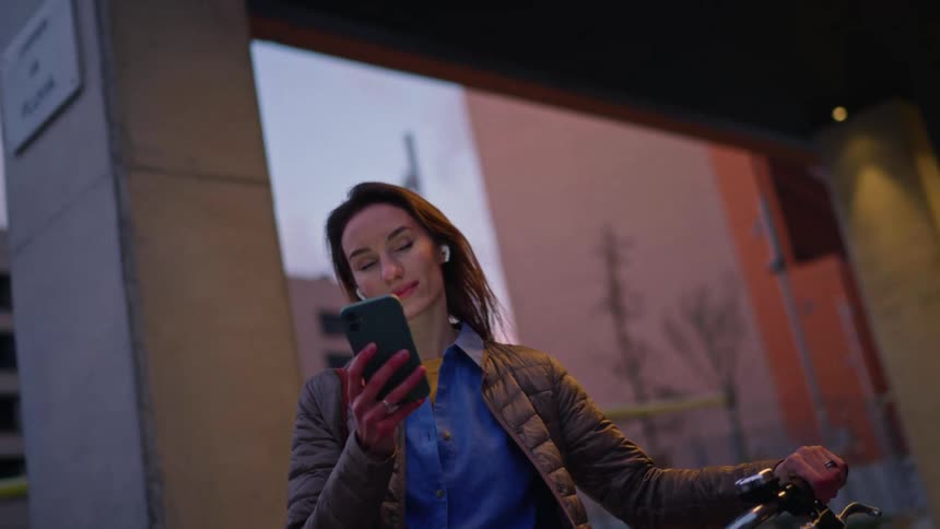 A woman holds a smartphone while standing beside a bicycle in an urban area at dusk